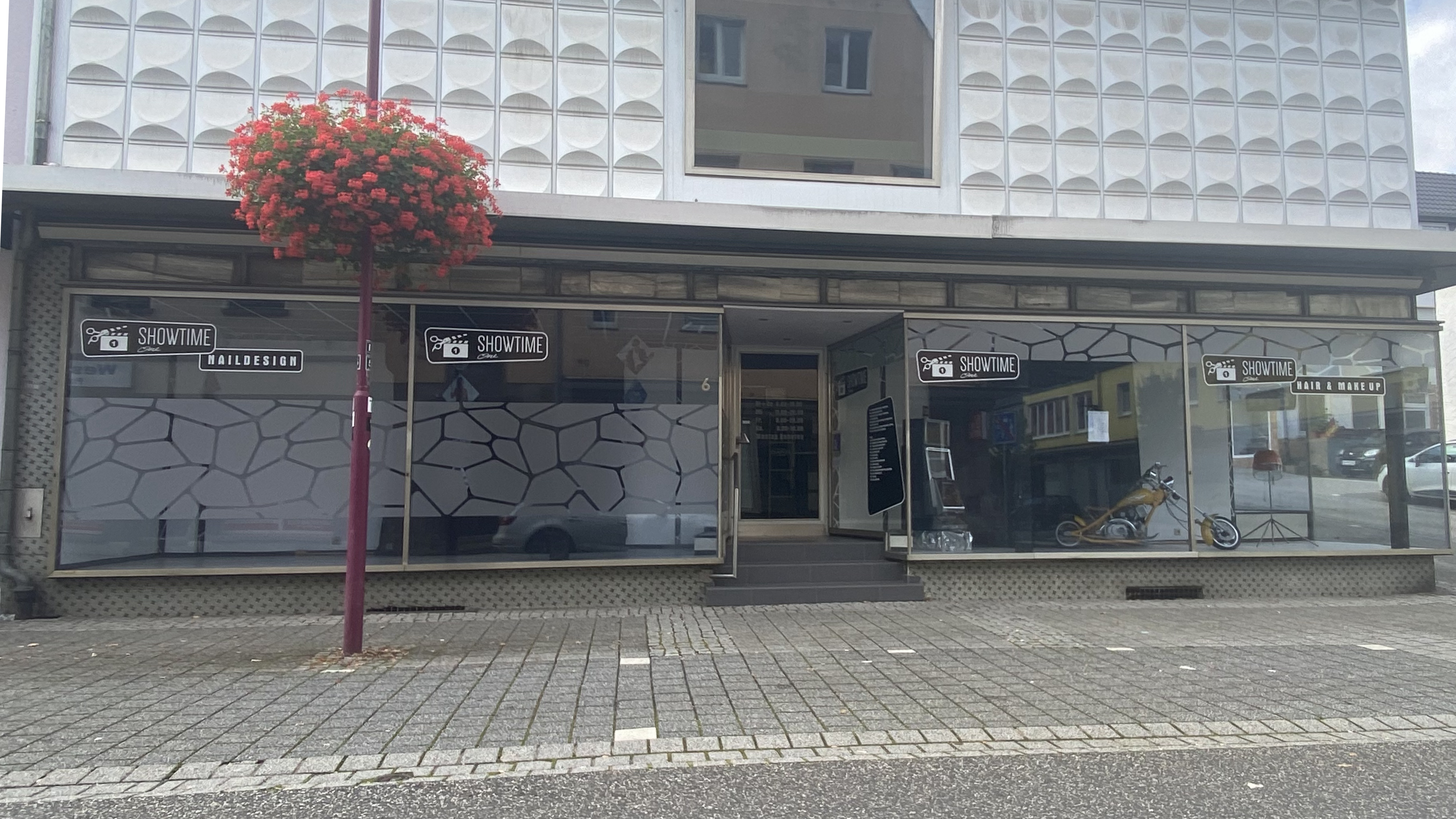 Aussenansicht des 'Showtime' Schönheitssalons in Westerburg, der Nageldesign und Haar- & Make-up-Services anbietet, mit Glasfenstern mit geometrischen Mustern, einem hängenden roten Blumenkorb und einer Motorrad-Dekoration im Inneren.