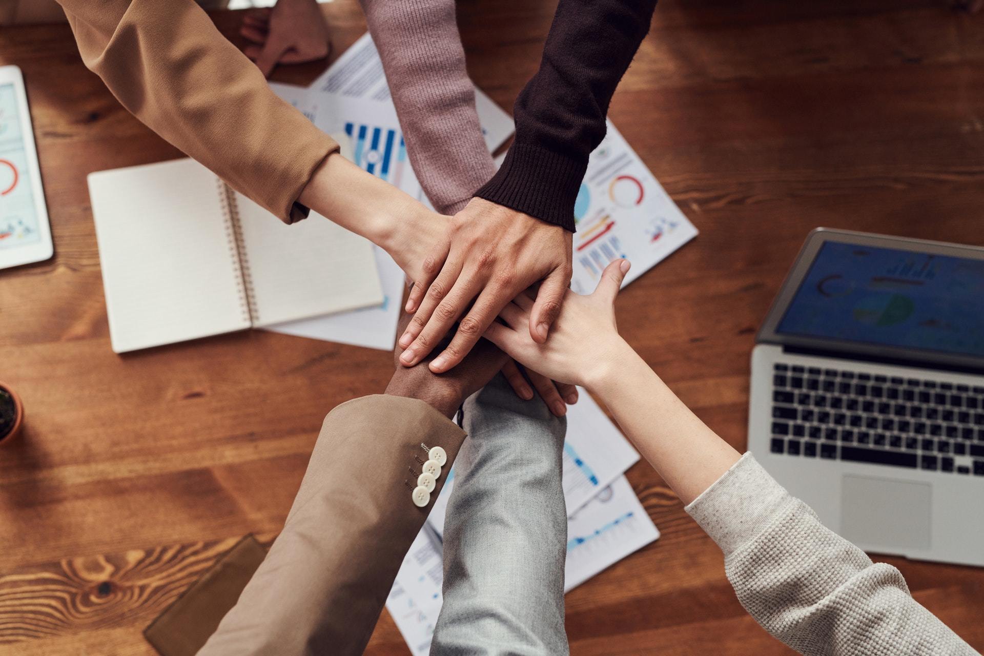 Übereinandergelegte Hände mehrerer Personen auf einem Holztisch mit Geschäftsunterlagen, Laptop und Notizbuch – Symbol für Teamwork und Zusammenarbeit regionaler Unternehmen im Westerburger Land.