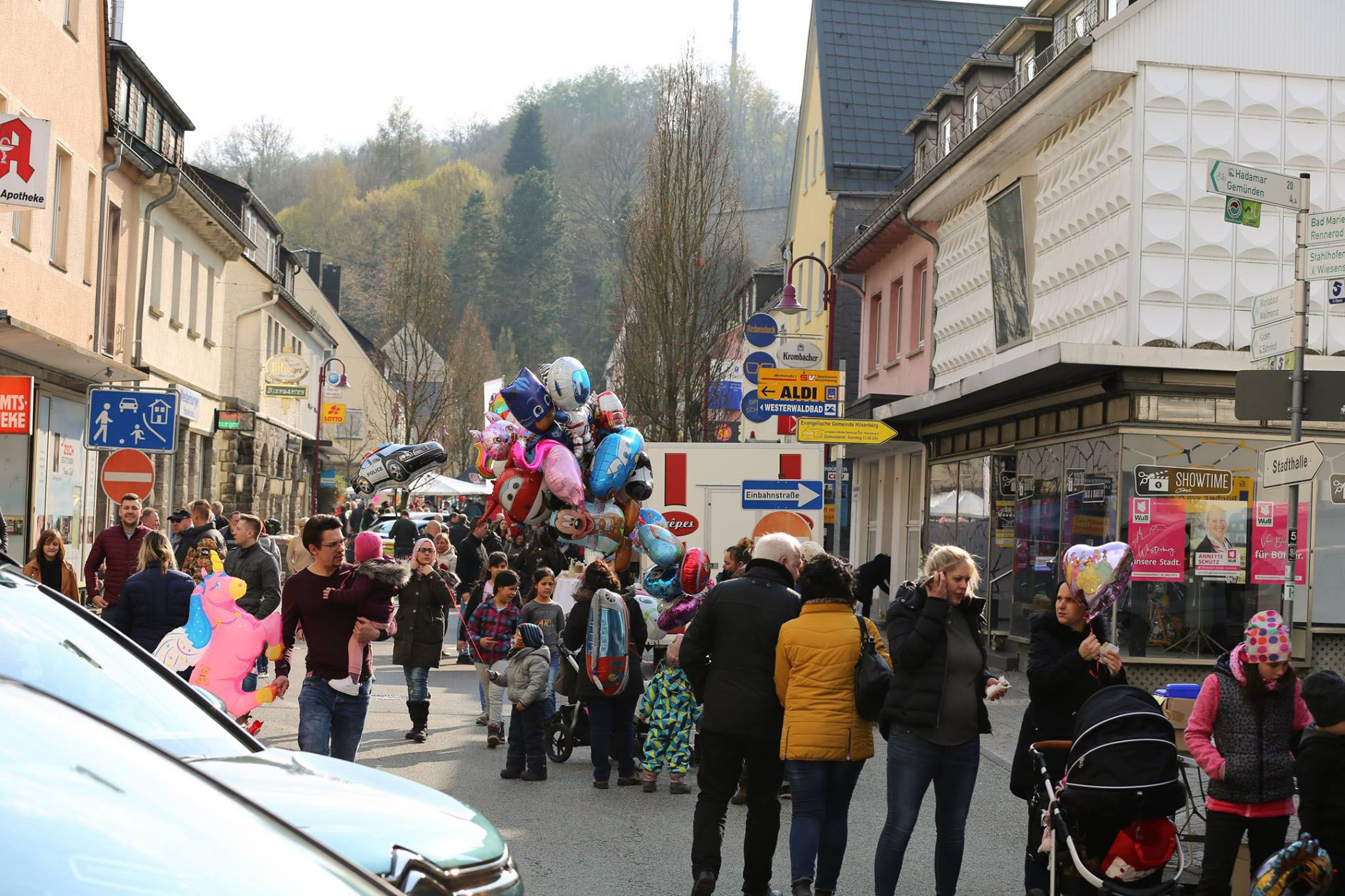 Bunte Szene auf einer belebten Straße in Westerburg: Familien, Besucher und Händler bei einem lokalen Event mit Luftballons, Geschäften (u.a. Apotheke, ALDI) und Schildern für Westerwaldbad, Hadamar – typisch für die Aktivitäten des Gewerbevereins Westerburger Land zur Stärkung des regionalen Wirtschaftsstandorts.
