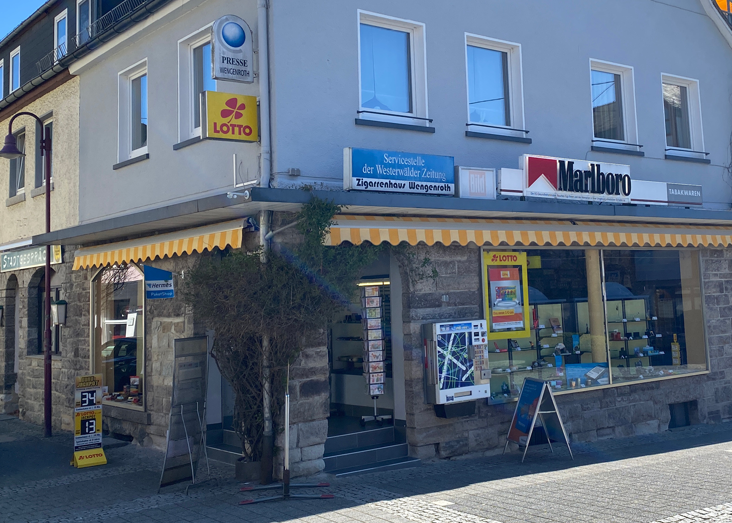 Aussenansicht des Zigarrenhaus Wengenroth, ein lokales Tabak- und Lotteriegeschäfts in Westerburg, mit Schildern für Lotto, Marlboro und die Westerwalder Zeitung-Servicestelle, gelb-weißem Vordach und Steinfassade.