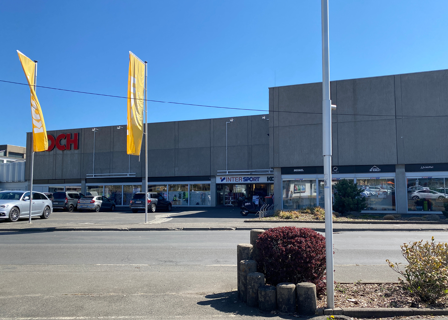 Kommerzielles Gebäude in Westerburg mit den Geschäften 'Koch' und 'INTERSPORT', gelben Fahnen, geparkten Autos und klarem Himmel – Teil des lokalen Gewerbeviertels, das vom Gewerbeverein Westerburger Land gefördert wird.