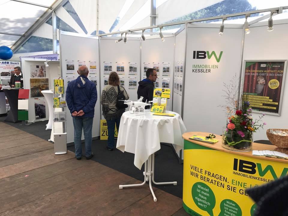 Bild des IBW Immobilien Kessler-Stands auf der Hui Wäller Messe, einer regionalen Veranstaltung des Gewerbevereins Westerburg, mit Besuchern, die sich an Informationswänden orientieren und Promotionsmaterialien auf einem Tisch betrachten.