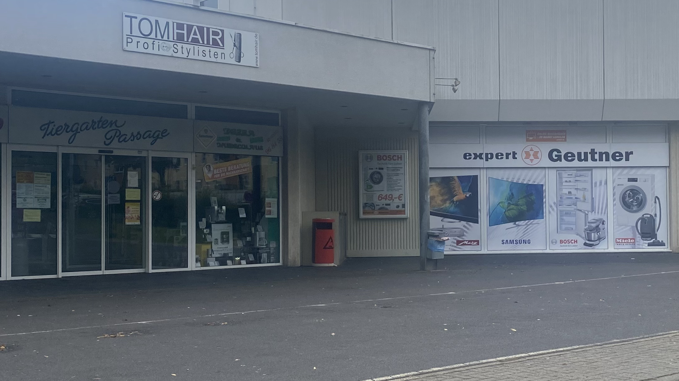 Ansicht von Geschäftsfassaden in Westerburg: Links 'TOMHAIR Profi Stylisten' über 'Tiergarten Passage', rechts 'expert Geutner' (Elektro- und Haushaltsgeräte mit Marken wie Samsung, Bosch, Miele), repräsentiert die lokale Wirtschaft, die der Gewerbeverein Westerburg fördert.