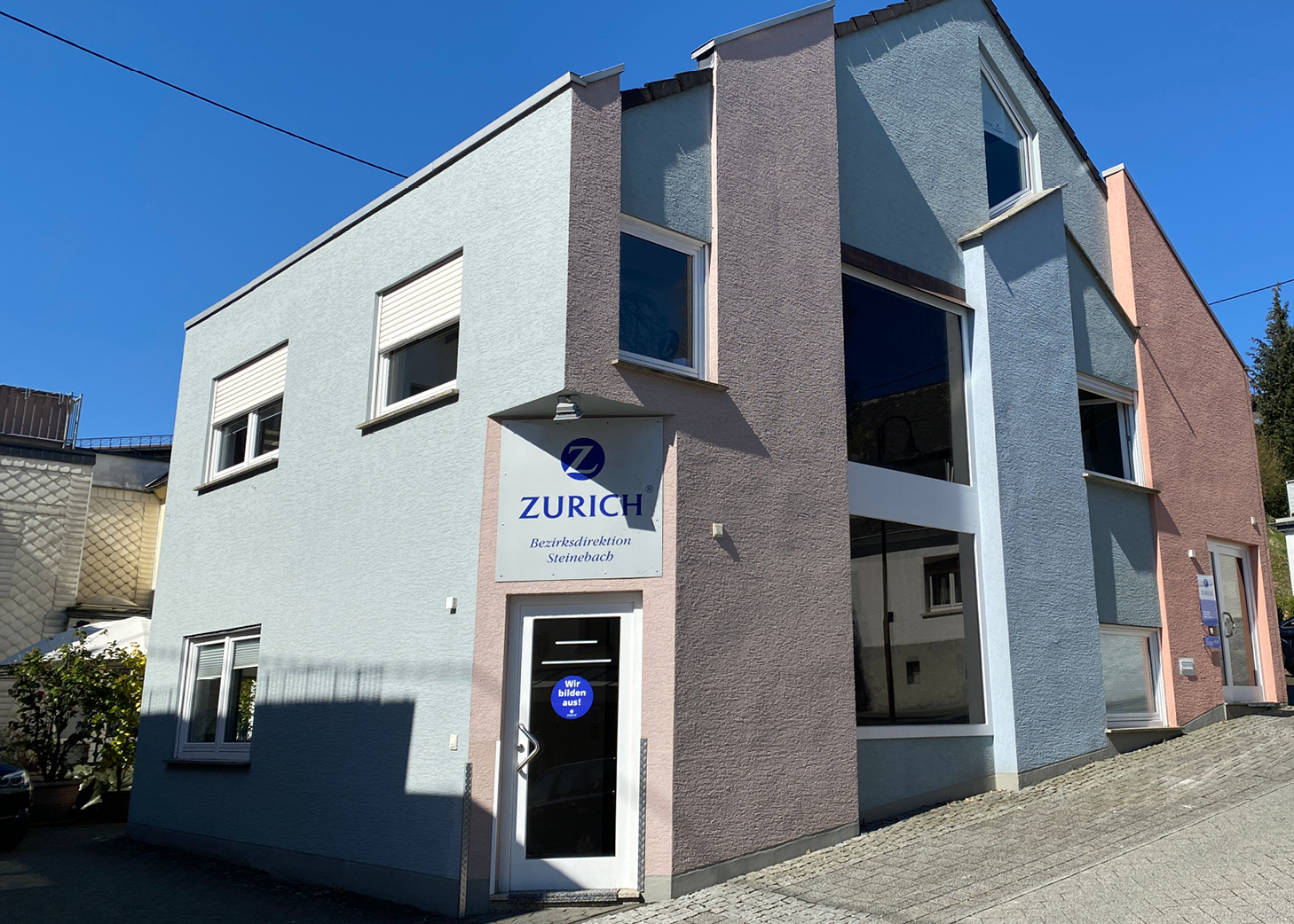 Außenansicht des modernen Bürogebäudes der Zurich Bezirksdirektion Steinebach, mit sichtbarem Firmenlogo und Türschild.