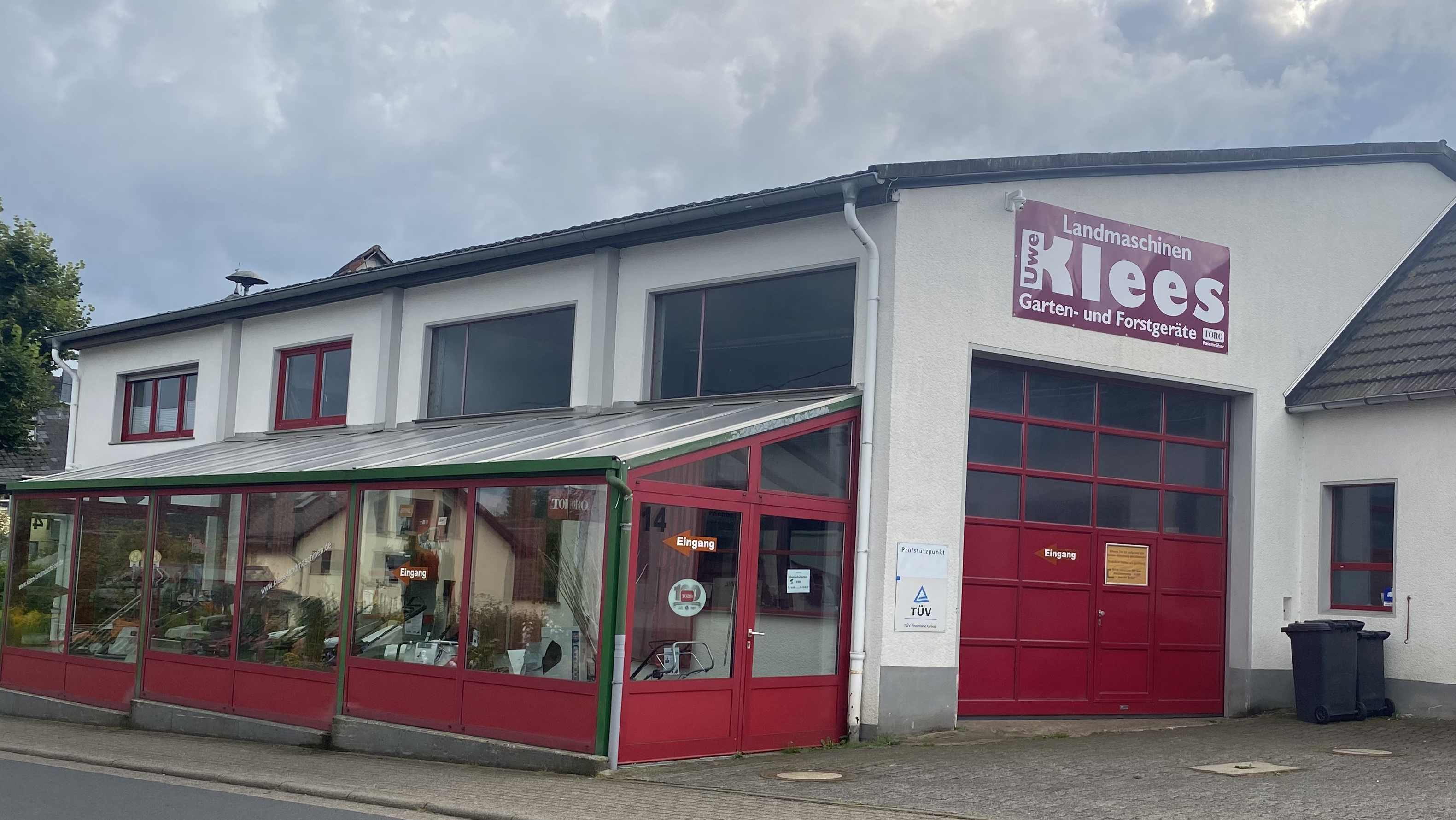 Außenansicht des Unternehmens Uwe Klees Landmaschinen in Westerburg, das Garten- und Forstgeräte sowie Landmaschinen anbietet, mit roten Türen, Fenstern und einem Schild, das die Unternehmensbezeichnung und Produkte zeigt.