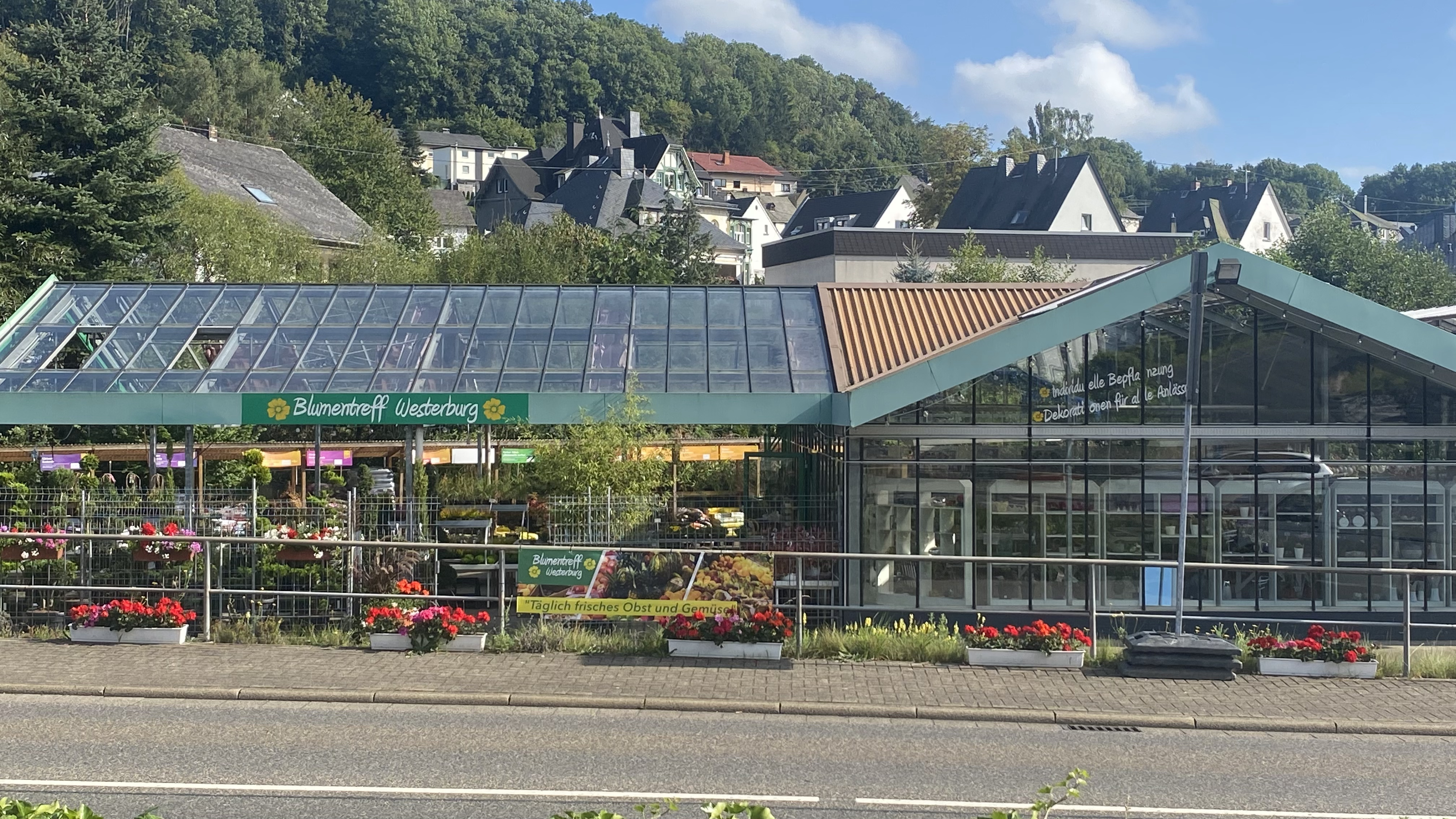Blumentreff Westerburg – Blumenladen mit Gewächshaus und bunten Blumen vor Häusern und Hügeln in Westerburg, repräsentiert lokale Unternehmen des Gewerbevereins Westerburger Land.