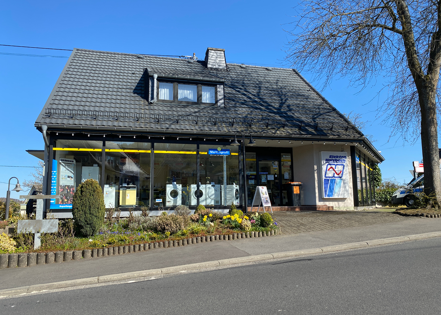Schaufenster von Elektro Holzbach, einem lokalen Elektrofachgeschäft in Westerburg, mit großen Fenstern, in denen Geräte ausgestellt sind, einem gepflegten Garten und einem strahlend blauen Himmel – ein Beispiel für die regionalen Unternehmen, die vom Gewerbeverein Westerburg gefördert werden.