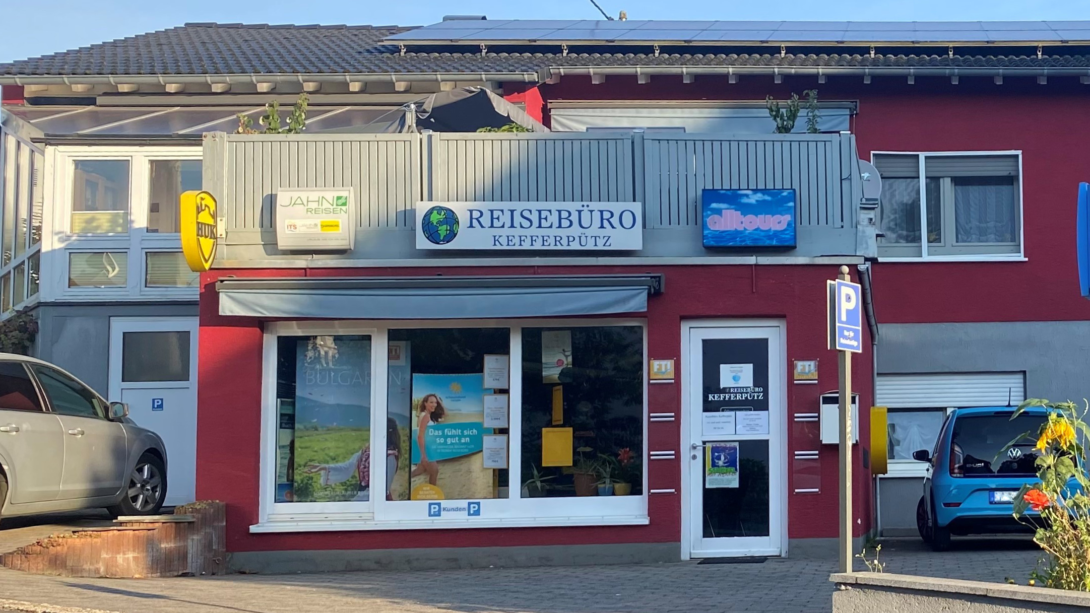 Fassade des Reisebüros Kefferputz in Westerburg: Rotes Gebäude mit Schildern für Reisebüro Kefferputz, Jahn Reisen, alltours und HUK, sowie Kundenparkplätzen.