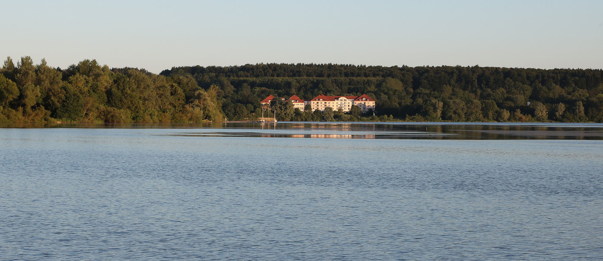 Klare Wasserfläche eines Sees mit bewaldeter Uferlinie und einem Gebäude mit roten Dächern im Hintergrund, charakteristisch für die Landschaft der Verbandsgemeinde Westerburg.