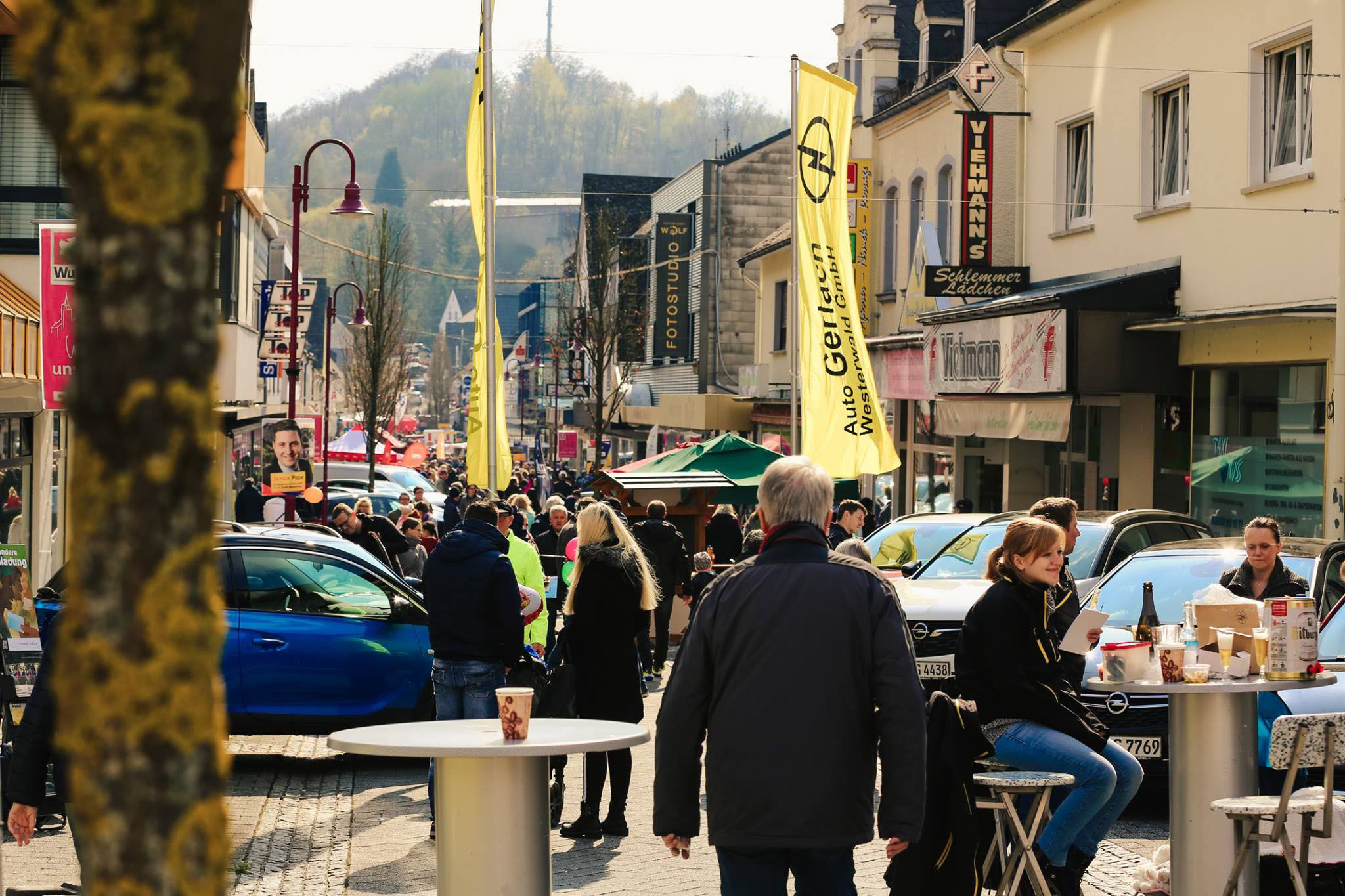 Buntes Straßenfest oder verkaufsoffener Sonntag in Westerburg: Menschen bevölkern die Fußgängerzone mit lokalen Geschäften, Outdoor-Sitzgelegenheiten und gelben Werbefahnen, die das lebendige Gewerbeleben der Region zeigen.