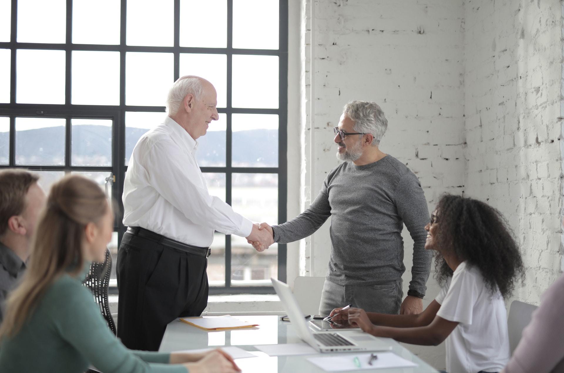 Zwei Unternehmer schütteln sich bei einem Geschäftsmeeting im Büro die Hände, während Kollegen an einem Tisch mit Laptops und Dokumenten zusammensitzen – ein Symbol für die Zusammenarbeit, die der Gewerbeverein Westerburg zur Stärkung des regionalen Wirtschaftsstandorts fördert.
