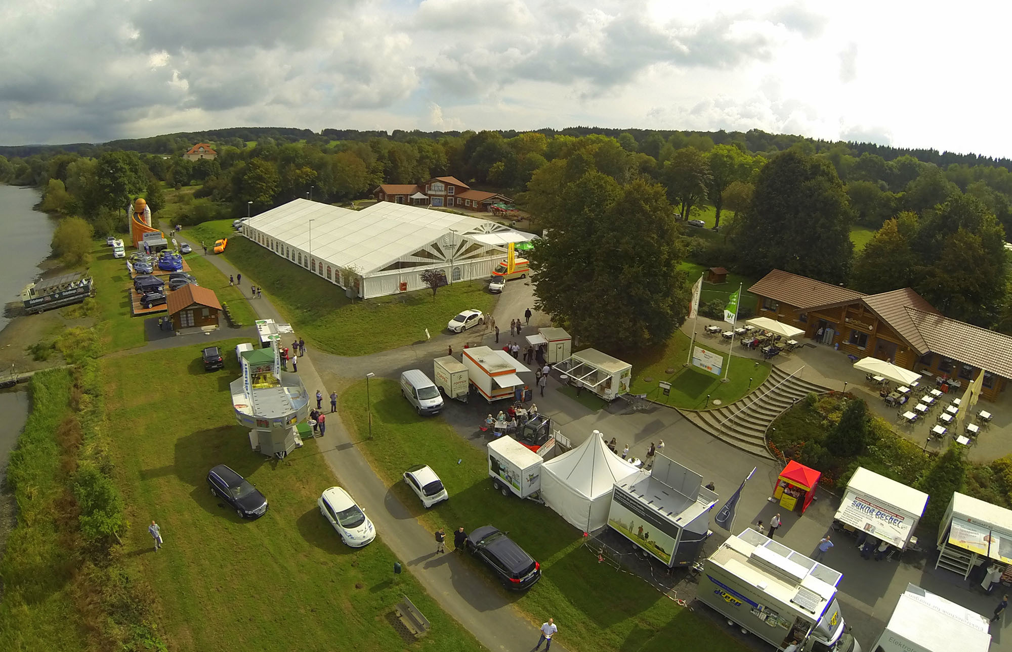 Luftaufnahme der Hui Wäller Messe des Gewerbevereins Westerburg: Zelte, Stände, Besucher und eine malerische Seenlandschaft mit Bäumen und Gebäuden.