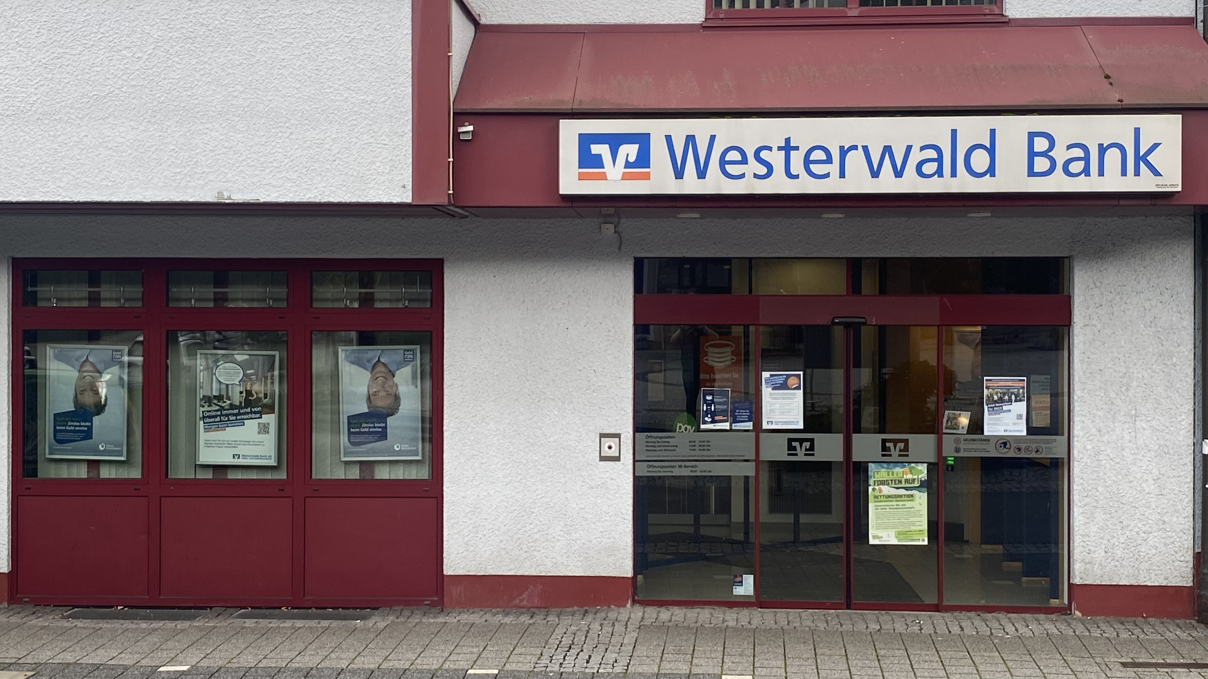 Westerwald Bank Filiale in Westerburg mit roter und weißer Fassade, Glasschiebetüren und Werbeplakaten im Schaufenster.